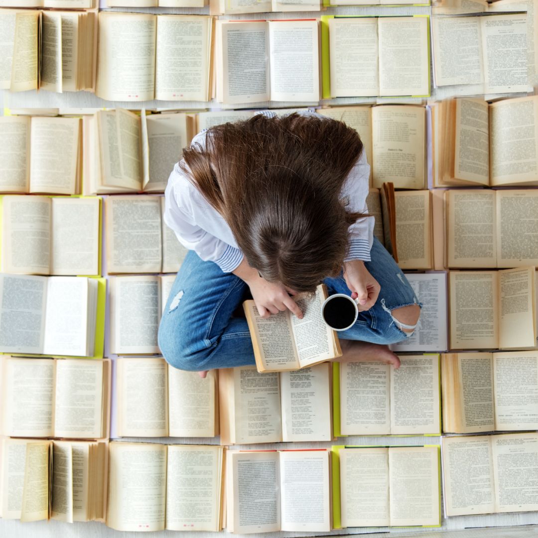 Image of woman sat on a floor of open books, reading a book an drinking a drink