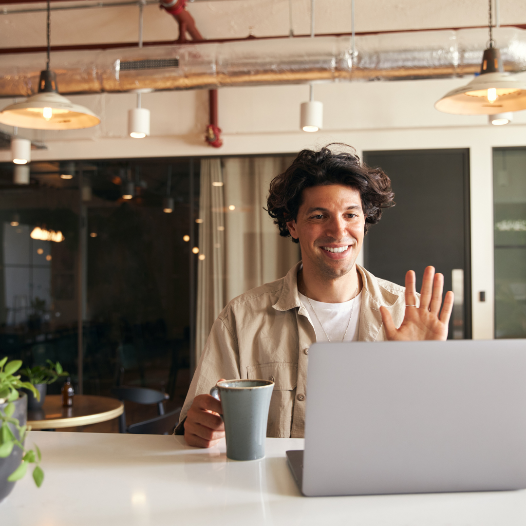 Image of man sat at table with a drink on a video call waving to laptop screen