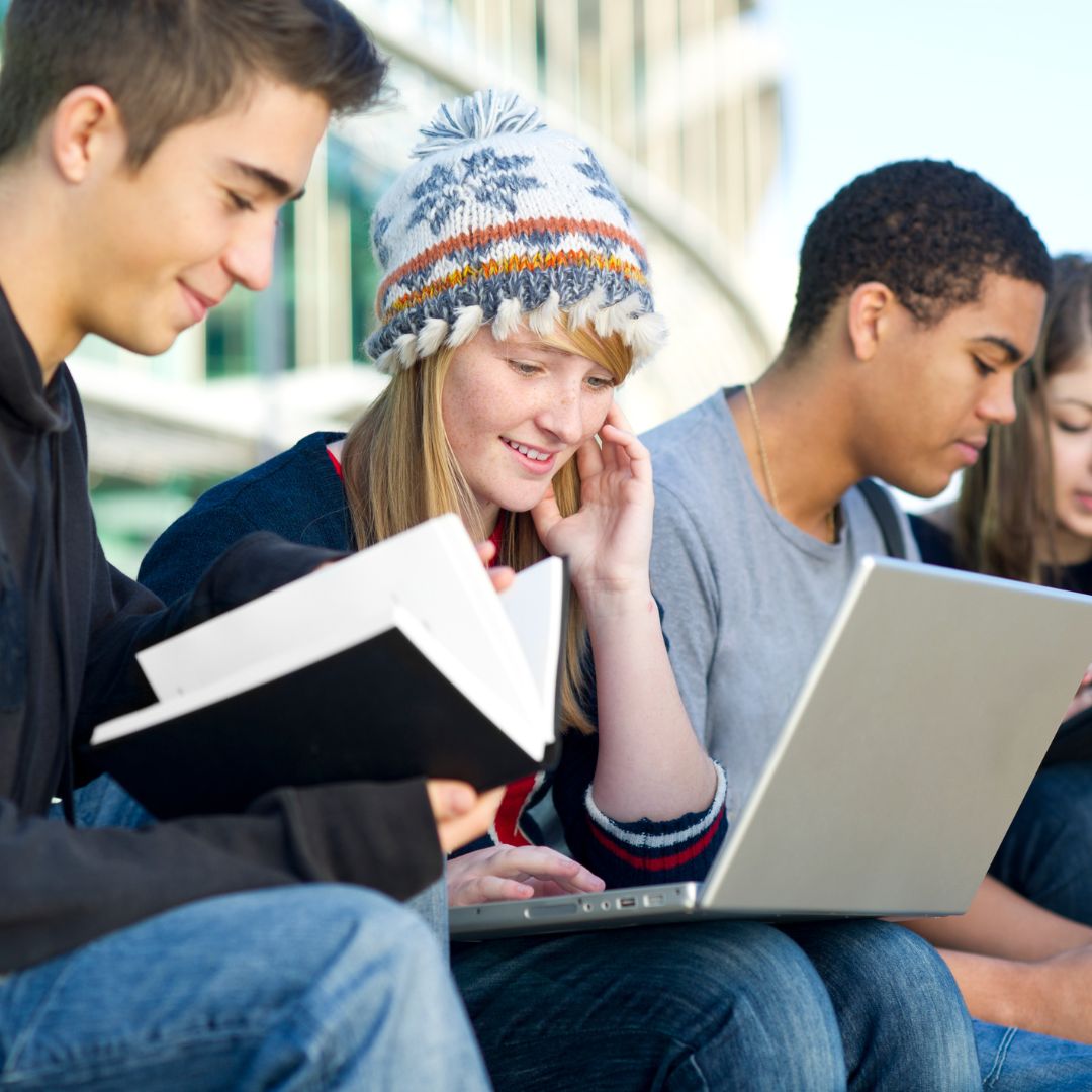 Image of students sat on steps to university building looking at books and laptop