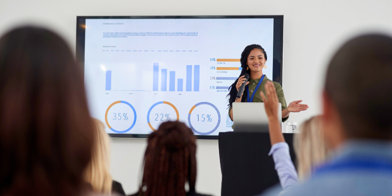 image of woman presenting, board behind her showing graphs