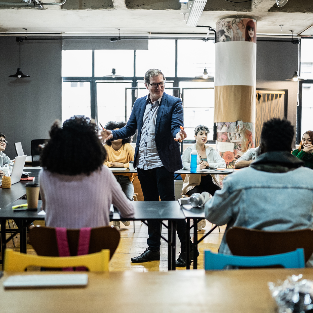 image of lecturer stood in middle of classroom surrounded by students teaching