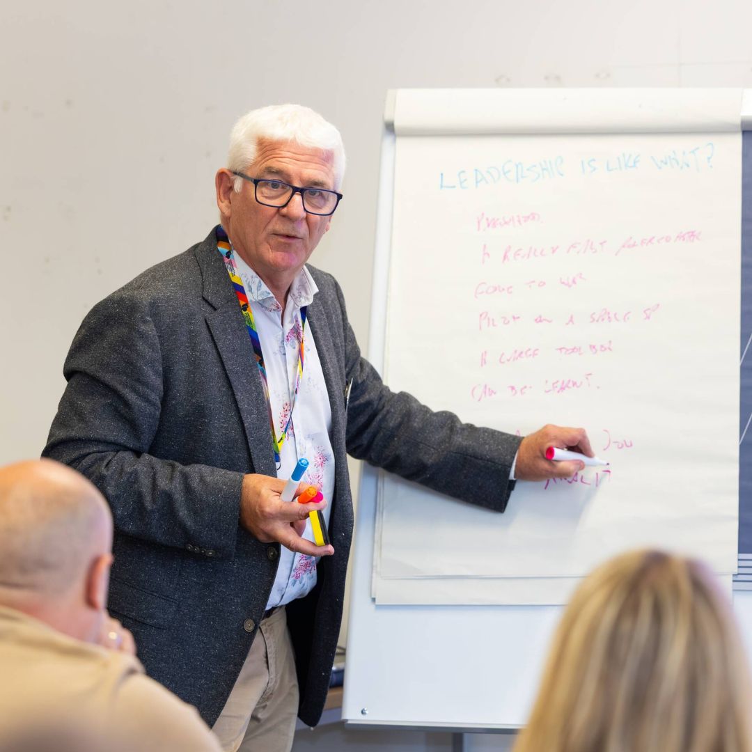 Image shows a man writing on a board and discussing with people in the room.