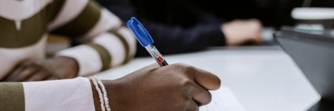 A student writing with a blue pen and wearing a green striped jumper