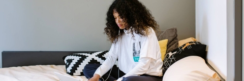 Image shows student on bed with headphones plugged in while using a laptop.